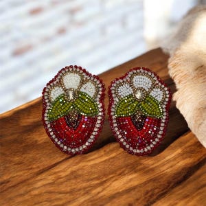 May include: A pair of beaded strawberry earrings. Each earring has a red, green, and white strawberry design with a beaded border. The earrings are displayed on a wooden surface.