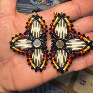 Peut inclure: Une paire de boucles d'oreilles faites à la main avec un motif floral. Les boucles d'oreilles sont ornées de perles noires, rouges, jaunes et blanches, entourant un motif central de piquants crème et marron. Chaque boucle d'oreille a un petit centre rond argenté.