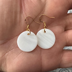 May include: A pair of white shell earrings with gold hooks. The earrings are round and have a slightly textured surface.