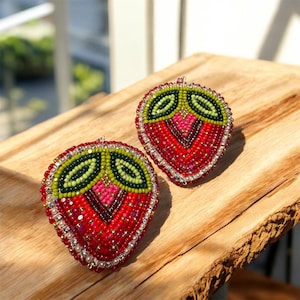 May include: Two beaded strawberry brooches. The strawberries are red with green leaves and a pink center. The brooches are embellished with small beads and pearls, and are set on a wooden surface.