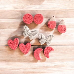 May include: An assortment of decorative stamps in various shapes, including circles, teardrops, hearts, butterflies, and crescents. The stamps are in shades of red and gray, with detailed floral patterns. The stamps are arranged on a light wooden surface.