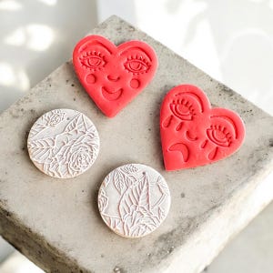 May include: Four decorative pieces: two red heart-shaped and two white circular. The hearts have faces with eyes and smiles or tears. The circles feature floral and leaf patterns, all on a gray surface.