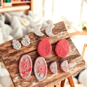 May include: A collection of decorative, textured plaster shapes in various designs. Includes red egg shapes with floral patterns, gray crescent moons, and oval shapes with hearts and flowers. The items are arranged on a rustic wooden surface.