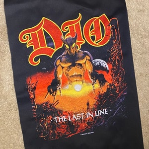 May include: Black patch with a red and yellow image of a horned, muscular figure with a chain in each hand. The figure stands in front of a fiery, apocalyptic scene with a large moon in the background. The text "DIO" is in a large, stylized font at the top of the patch. The text "THE LAST IN LINE" is at the bottom of the patch.