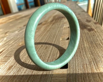 May include: A light green jade bangle bracelet. The bangle is smooth and polished, and it has a slightly oval shape.
