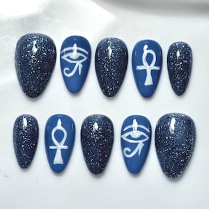 May include: A set of ten press-on nails in a mix of dark blue glitter and solid blue finishes. Some nails feature hand-painted white Egyptian symbols, specifically the Eye of Horus and the Ankh, arranged in a rounded almond shape design.