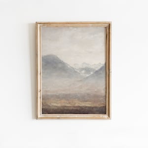 May include: A framed painting of a misty mountain landscape with a light brown frame. The painting depicts a mountain range with a hazy sky and a foreground of brown and green.