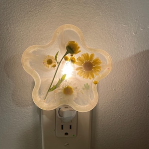 May include: A star-shaped night light with a clear resin design, featuring yellow and white daisies. The night light is plugged into a white electrical outlet, illuminating the flowers. The overall design is a floral theme.