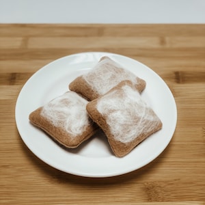 May include: Three brown felt pillows with white fluffy accents are arranged on a white plate. The plate is on a wooden surface.