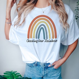 以下が含まれることがあります： 虹のグラフィックと「Chasing Our Rainbow」というテキストが書かれた白いTシャツです。