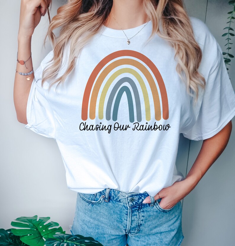 以下が含まれることがあります： 虹のグラフィックと「Chasing Our Rainbow」というテキストが書かれた白いTシャツです。