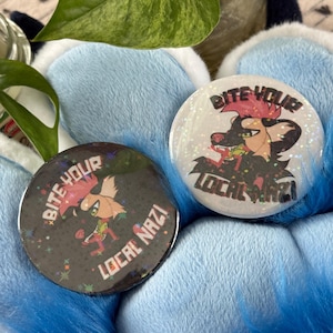 May include: Two circular badges with the text "Bite Your Local Nazi". One is black with a cartoon design, the other white. Both have a glittery finish. The badges are set against a blue background.