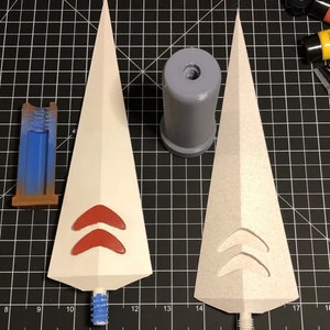 Puede incluir: Dos puntas de lanza de espuma blanca con marcas pintadas de rojo. Las puntas están unidas a bases de plástico gris. Las bases tienen un agujero roscado en la parte superior.