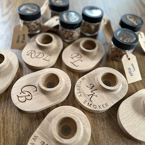 Puede incluir: Varios ahumadores de whisky de madera con iniciales grabadas y la inscripción "Whiskey Smoker". También hay pequeños frascos de vidrio llenos de virutas de madera y etiquetados como "Apple Wood". Los artículos están dispuestos sobre una superficie de madera.