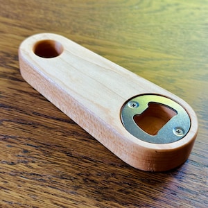 May include: A wooden bottle opener with a metal cap opener. The opener is light brown and has a smooth finish.