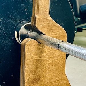 May include: A wooden barbell safety stand with a brown stain finish. The stand is designed to hold a barbell in place during exercises.