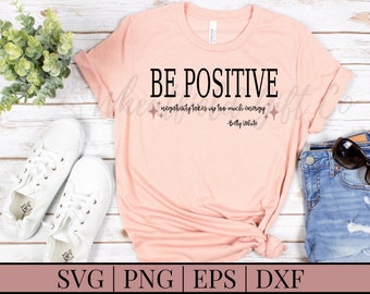May include: A pink t-shirt with the text "BE POSITIVE" in black. The text "negativity takes up too much energy - Betty White" is also printed in black on the shirt. The shirt is on a white wooden surface with a pair of white trainers, a pair of blue jeans, and a pair of sunglasses.