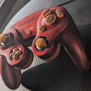May include: A red Nintendo GameCube controller with gold buttons and a "START/PAUSE" button label. The controller has a textured surface and is resting on a black surface.