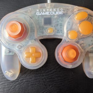 May include: A clear, translucent Nintendo GameCube controller with orange buttons. The controller has a clear plastic shell and orange buttons, including the control stick, directional pad, and face buttons. The text "NINTENDO GAMECUBE" is printed on the top of the controller.