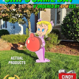 May include: A handcrafted wooden yard sign featuring Cindy Lou Who from the film How the Grinch Stole Christmas. The sign is painted in pink, red, and white and features a cartoon image of Cindy Lou Who holding a red ball. The sign is surrounded by green grass and a house in the background. The text "HUGE SALE SHIPS SAME DAY IF ORDERED BY 4PM EST." is at the top of the image. The text "ACTUAL PRODUCTS" is below the sign. The text "5 HANDCRAFTED YARD SIGNS" is below the sign. The text "CINDY LOU WHO" is in a red square below the sign.