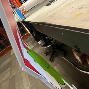 May include: A close-up of a large, printed sign featuring a green and white cartoon character with a red and white striped background. The sign is being prepared for display.