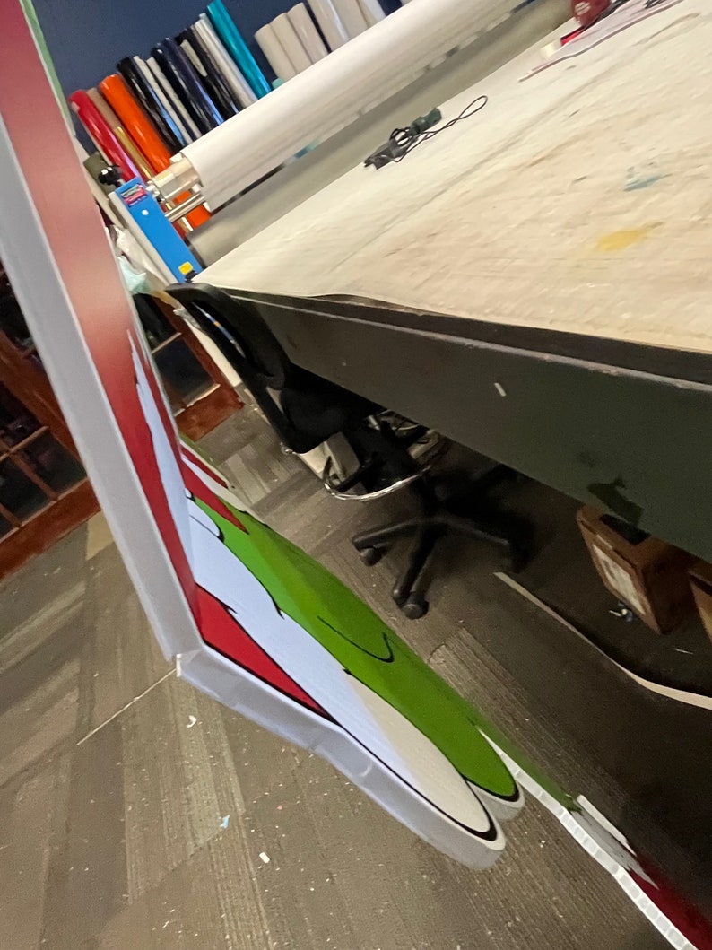 May include: A close-up of a large, printed sign featuring a green and white cartoon character with a red and white striped background. The sign is being prepared for display.