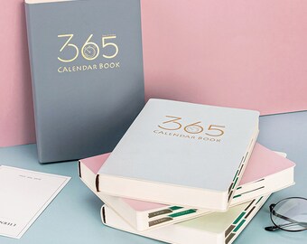May include: A stack of 365 Calendar Books in various colors, including gray, light blue, and pink. The books have gold lettering and a clock graphic. The books are on a blue surface with a pink background.