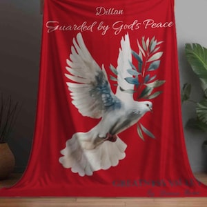 May include: A red blanket featuring a white dove with blue and gray accents, holding a green olive branch. The blanket has the text "Dillon Guarded by God's Peace" in white script. Decorative home decor.