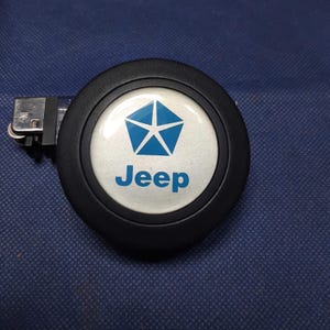 May include: A black Jeep lighter with a white and blue logo. The logo features a blue pentagon star and the word "Jeep" in blue. The lighter is on a blue surface.