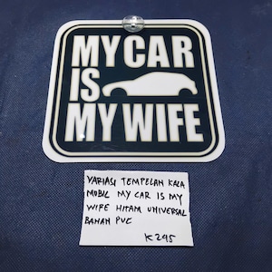 May include: A white and black sticker with the text "MY CAR IS MY WIFE" and a car silhouette. The sticker is rectangular with rounded corners and is attached to a blue surface. Additional text is present at the bottom.