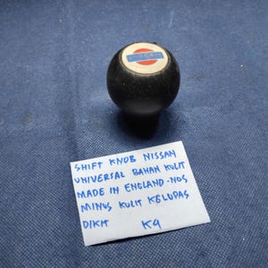 May include: A black shift knob with a white, red, and blue emblem. The knob has a rounded shape and is likely for a vehicle's gear shift. The text on the attached paper reads "SHIFT KNOB NISSAN".