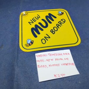 Op de afbeelding: Een gele rechthoekige plaat met afgeronde hoeken, met de tekst "NEW MUM ON BOARD" in blauw en twee cartoonbijen. Een zuignap is bevestigd. Een handgeschreven notitie staat eronder.