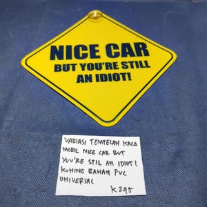 May include: A yellow diamond-shaped sign with the text "NICE CAR BUT YOU'RE STILL AN IDIOT!" in bold, dark blue letters. The sign has a small gold-colored attachment at the top. The sign is on a blue surface.