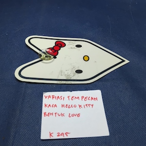 May include: White, heart-shaped Hello Kitty sticker with black outline, red bow, and yellow and black accents. A small, clear, round object is attached to the sticker. The text on the paper reads: "VARIASI TEM PELAN KACA HELLO KITTY BENTUK LOVE K 295."