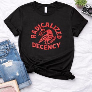Könnte beinhalten: Schwarzes T-Shirt mit roter Grafik. Die Grafik zeigt einen Vogel mit einem Zweig in einem kreisförmigen Design, mit den Worten "RADICALIZED BY BASIC DECENCY" in fetter, serifenloser Schrift. Das Shirt wird mit Jeans und einem Buch gestylt.