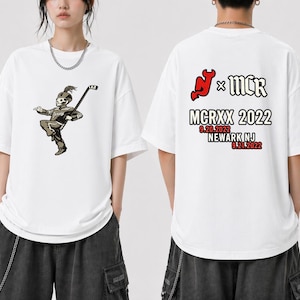 Könnte beinhalten: Weißes T-Shirt mit einem Skelett-Marschkapellenmitglied, das einen Hockeyschläger hält. Auf der Rückseite des Shirts steht der Text "MCRXX 2022" mit Veranstaltungsort und Datum.