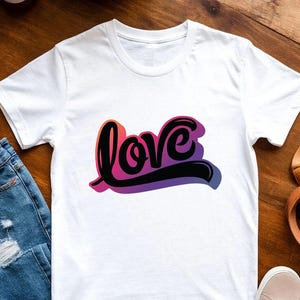 May include: White t-shirt with the word "love" in a gradient of pink, purple, and black. The word is in a cursive font with a shadow effect. The shirt is displayed on a wooden surface with jeans and sneakers.