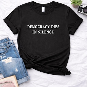 Pode incluir: T-shirt preto com o texto branco "DEMOCRACY DIES IN SILENCE". A camisa é combinada com jeans azuis e um livro em um lençol branco. Uma pequena planta e uma câmera também são visíveis.