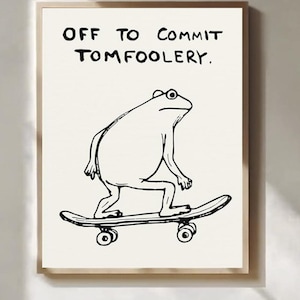 Op de afbeelding: Een ingelijste print met een zwart-wit illustratie van een kikker op een skateboard. De tekst boven de kikker luidt "OFF TO COMMIT TOMFOOLERY."