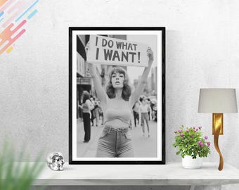 May include: Black and white photograph of a woman holding a sign that reads "I do what I want!" The woman is wearing a crop top and jeans. The photo is framed in a black frame.