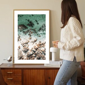 Könnte beinhalten: Ein gerahmter Kunstdruck mit einer Luftaufnahme einer felsigen Küstenlinie. Das Bild zeigt türkisfarbenes Wasser, weißen Sand und braune Felsen. Der Rahmen ist helles Holz, und das Kunstwerk wird auf einem Holzschrank ausgestellt.