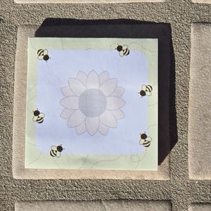 May include: A light blue notepad with a white sunflower and bees in black and yellow. The notepad has a green border with a dotted line pattern.