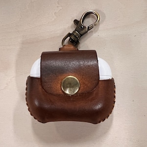 Puede incluir: Una funda de cuero marrón para AirPods con cierre de latón y botón. La funda está diseñada para guardar auriculares AirPods blancos. El cuero tiene un aspecto envejecido y las costuras son visibles en los bordes.