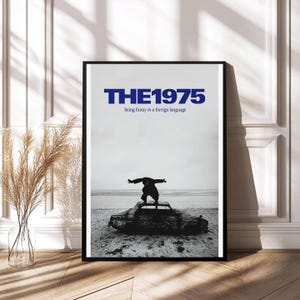 May include: Black framed poster featuring the band name "THE1975" in blue, with the text "being funny in a foreign language" below. The image shows a person standing on a car on a beach. The car is partially submerged in sand.