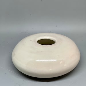 Può includere: Un vaso in ceramica bianca con una base rotonda e piatta e una piccola apertura nella parte superiore. Il vaso ha una finitura liscia e lucida.