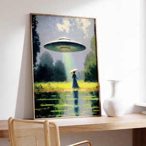 Op de afbeelding: Een ingelijst schilderij van een vrouw in een jurk en hoed die op een veld staat, met een vliegende schotel die boven haar zweeft. De schotel zendt een lichtstraal uit. Het schilderij is geplaatst tegen een witte muur.