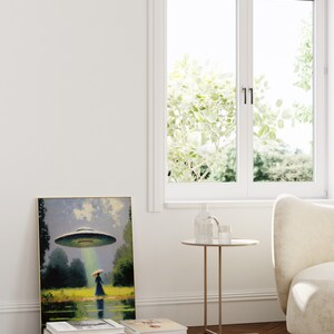 Op de afbeelding: Een ingelijst schilderij van een vrouw die in een veld staat met een vliegende schotel die boven haar zweeft. Het schilderij staat op een plank met een stapel boeken eronder. Een kleine ronde tafel met een glazen vaas en een glas water staat voor het schilderij.