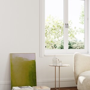 May include: A green and purple abstract painting in a gold frame leaning against a white wall. A stack of books is in front of the painting. A small round side table with a glass vase and a glass on top sits next to a white armchair.