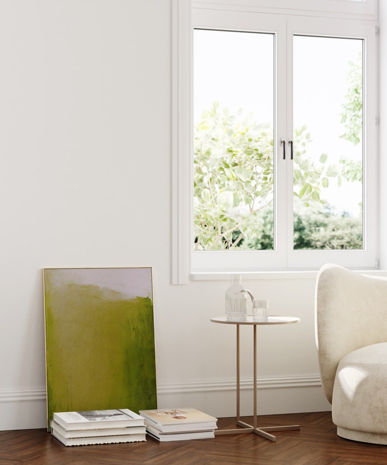 May include: A green and purple abstract painting in a gold frame leaning against a white wall. A stack of books is in front of the painting. A small round side table with a glass vase and a glass on top sits next to a white armchair.