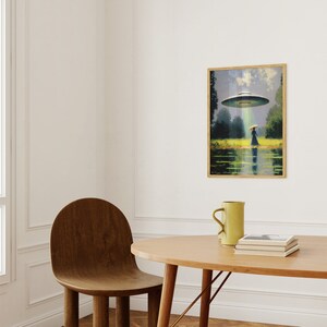 Op de afbeelding: Een ingelijst schilderij van een vrouw die over een veld loopt met een UFO die boven haar zweeft. Het schilderij is in een houten lijst en hangt aan een witte muur.